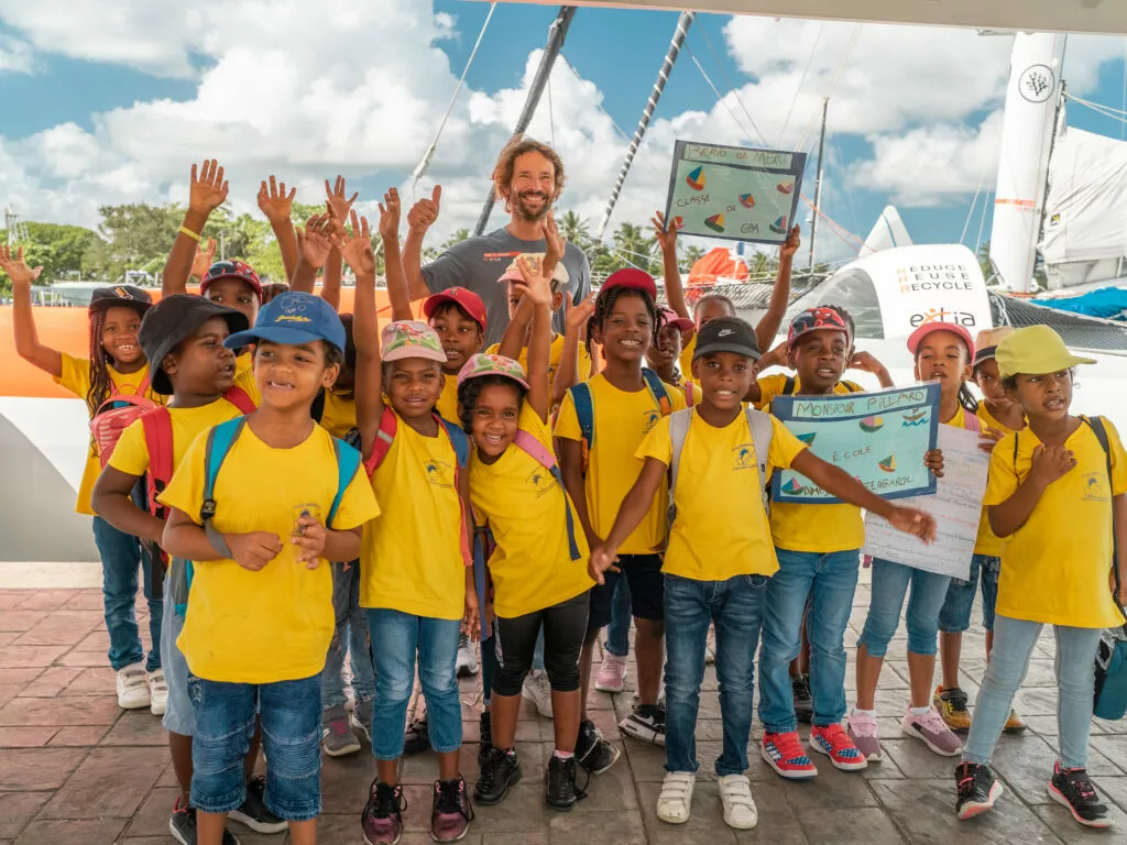 Un skipper entouré d'écoliers en tee-shirts jaunes, bras levés, sur le quai devant un trimaran